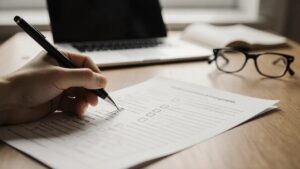 Hand füllt Versicherungs-Fragebogen mit Checkboxen aus, Laptop und Brille im Hintergrund